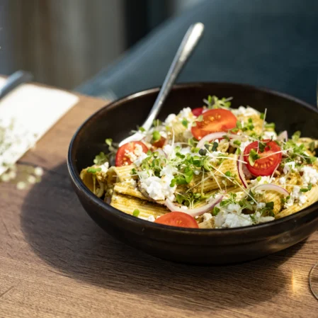 Delicious pasta salad with fresh tomatoes and a glass of white wine on a wooden dining table in a stylish hotel restaurant.