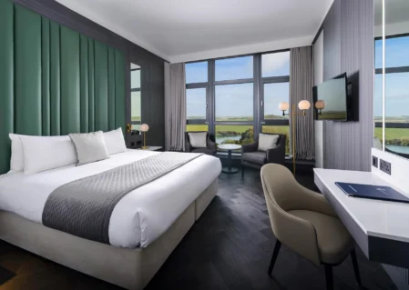 Modern hotel room with king-sized bed, scenic countryside view, large windows, cosy seating area, and flat-screen TV.