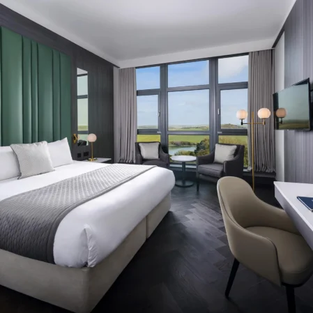 Modern hotel room with king-sized bed, scenic countryside view, large windows, cosy seating area, and flat-screen TV.