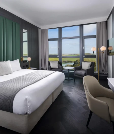 Modern hotel room with king-sized bed, scenic countryside view, large windows, cosy seating area, and flat-screen TV.