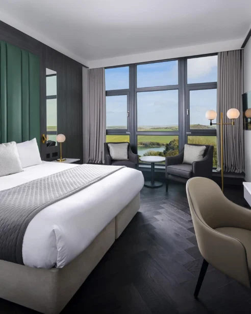 Modern hotel room with king-sized bed, scenic countryside view, large windows, cosy seating area, and flat-screen TV.