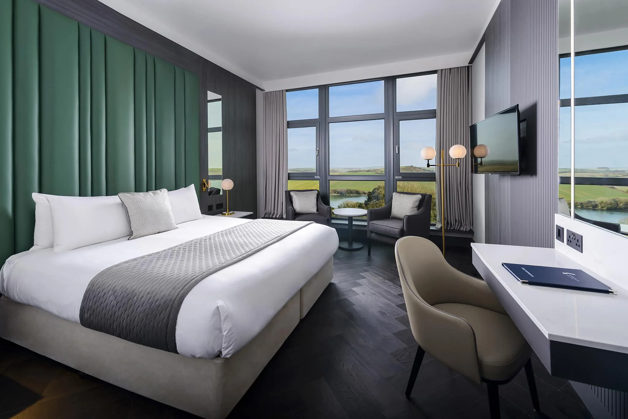 Modern hotel room with king-sized bed, scenic countryside view, large windows, cosy seating area, and flat-screen TV.