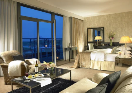 Elegant hotel suite with plush bedding, cosy seating area, city view, and wine service for a relaxing stay.