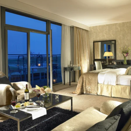 Elegant hotel suite with plush bedding, cosy seating area, city view, and wine service for a relaxing stay.