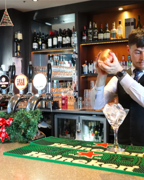 Bartender prepares cocktail at festive hotel bar with seasonal decor and variety of drinks on display.