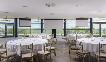 Spacious hotel event venue with round tables, panoramic countryside views, and elegant lighting for conferences or banquets.