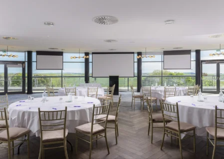 Spacious hotel event venue with round tables, panoramic countryside views, and elegant lighting for conferences or banquets.