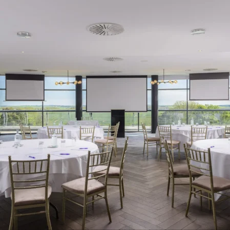 Spacious hotel event venue with round tables, panoramic countryside views, and elegant lighting for conferences or banquets.