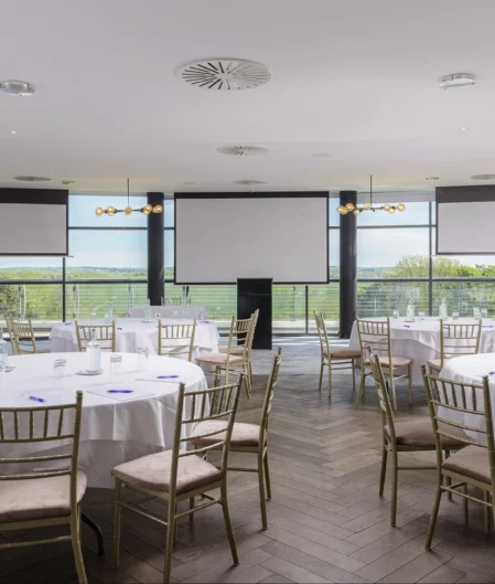 Spacious hotel event venue with round tables, panoramic countryside views, and elegant lighting for conferences or banquets.