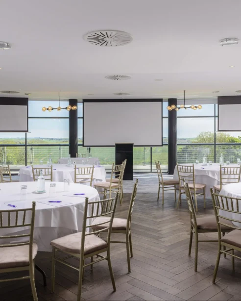 Spacious hotel event venue with round tables, panoramic countryside views, and elegant lighting for conferences or banquets.