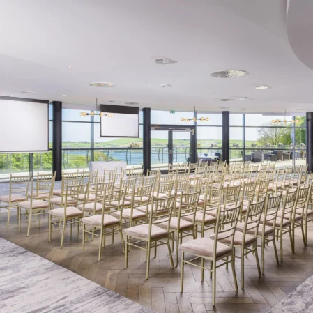 Elegant conference room with river views, gold chairs, and large projector screens at modern hotel venue.