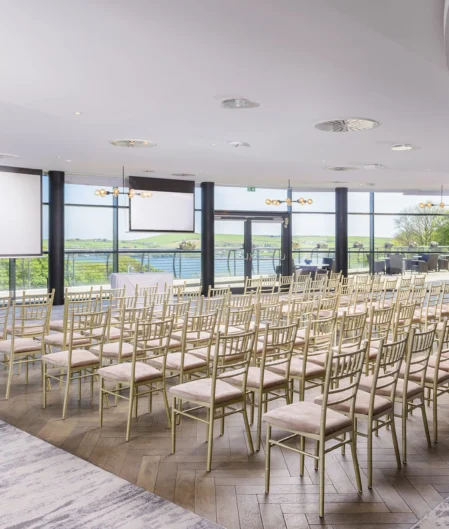 Elegant conference room with river views, gold chairs, and large projector screens at modern hotel venue.