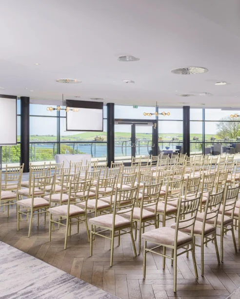 Elegant conference room with river views, gold chairs, and large projector screens at modern hotel venue.