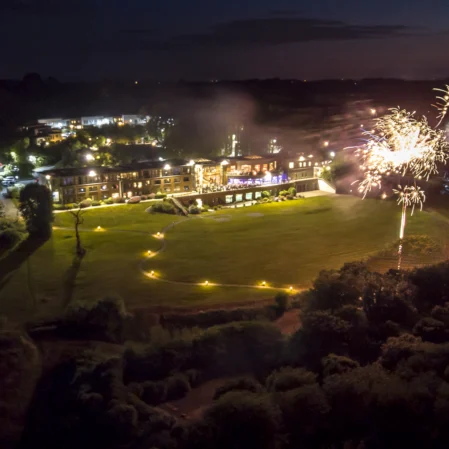Aerial view of hotel illuminated at night with spectacular fireworks display over lush gardens, offering a festive atmosphere.