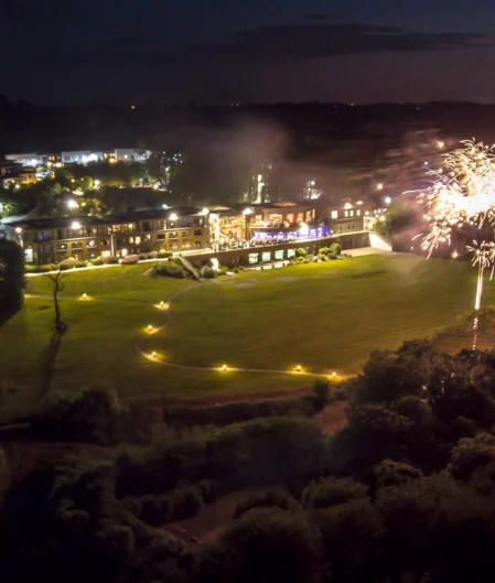 Aerial view of hotel illuminated at night with spectacular fireworks display over lush gardens, offering a festive atmosphere.