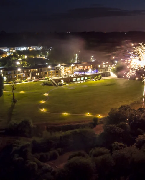 Aerial view of hotel illuminated at night with spectacular fireworks display over lush gardens, offering a festive atmosphere.