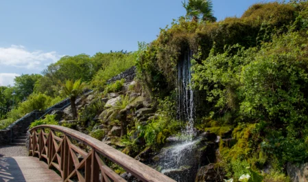 Lush hotel garden with waterfall, wooden walkway, bright flora, and serene atmosphere ideal for relaxation and nature walks.