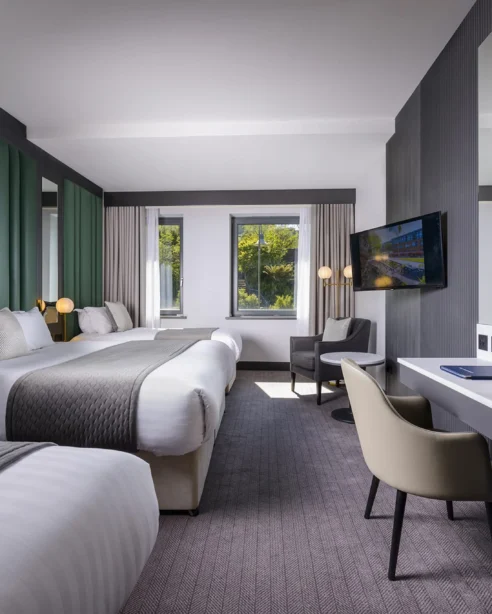 Modern hotel room with two beds, elegant decor, large windows, natural light, desk, and flat-screen TV for a cosy stay.