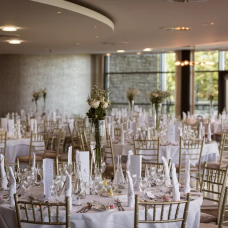 Elegant hotel banquet hall set for a wedding, featuring round tables with white linens, gold chairs, and floral centrepieces.