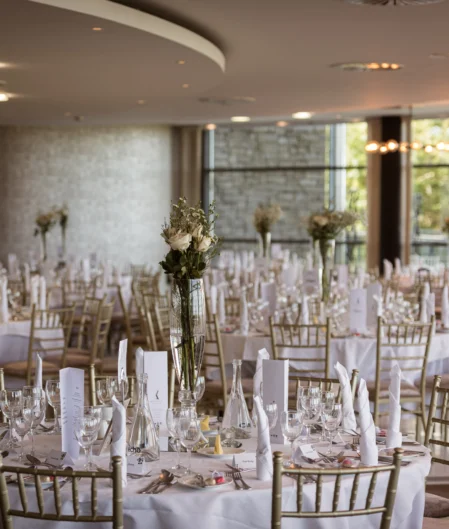 Elegant hotel banquet hall set for a wedding, featuring round tables with white linens, gold chairs, and floral centrepieces.