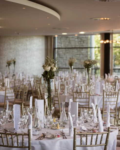 Elegant hotel banquet hall set for a wedding, featuring round tables with white linens, gold chairs, and floral centrepieces.
