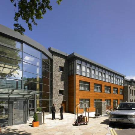 Modern hotel entrance with glass facade, welcoming staff, and elegant grey and wood accents under blue skies.