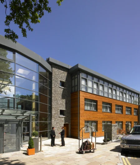 Modern hotel entrance with glass facade, welcoming staff, and elegant grey and wood accents under blue skies.