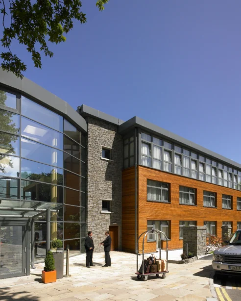 Modern hotel entrance with glass facade, welcoming staff, and elegant grey and wood accents under blue skies.