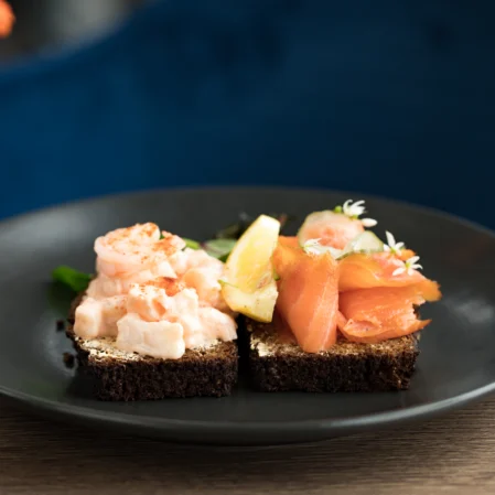 Gourmet seafood appetisers with prawns and smoked salmon on dark bread; wine glass and vibrant flowers in view.