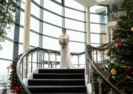 Bride on curved staircase near Christmas tree in elegant hotel lobby with large windows and festive decorations.