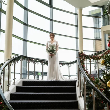 Bride on curved staircase near Christmas tree in elegant hotel lobby with large windows and festive decorations.