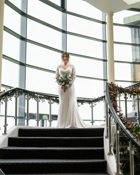 Bride on curved staircase near Christmas tree in elegant hotel lobby with large windows and festive decorations.