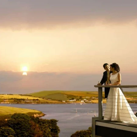 Couple enjoys romantic sunset view over river from hotel balcony, surrounded by lush green hills and serene waters.