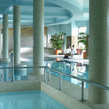 Indoor hotel pool with columns, lush plants, and guests relaxing near the water; tranquil and luxurious experience.