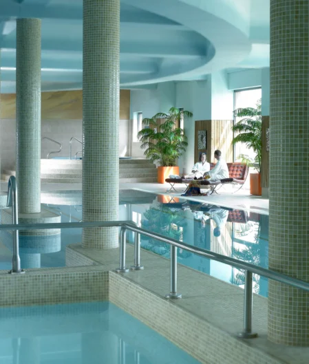 Indoor hotel pool with columns, lush plants, and guests relaxing near the water; tranquil and luxurious experience.