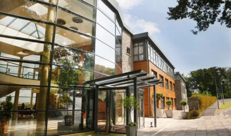 Modern hotel entrance with glass facade, outdoor plants, and welcoming path, showcasing contemporary architecture and inviting atmosphere.