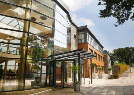 Modern hotel entrance with glass facade, outdoor plants, and welcoming path, showcasing contemporary architecture and inviting atmosphere.