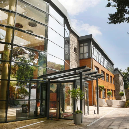 Modern hotel entrance with glass facade, outdoor plants, and welcoming path, showcasing contemporary architecture and inviting atmosphere.