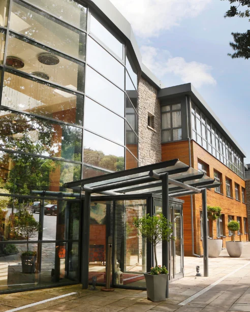 Modern hotel entrance with glass facade, outdoor plants, and welcoming path, showcasing contemporary architecture and inviting atmosphere.