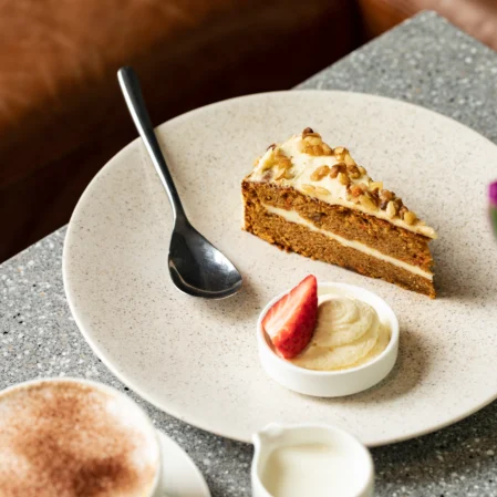 Slice of carrot cake with cream and strawberry on a speckled table, cosy atmosphere. Enjoy our café's delightful treats.