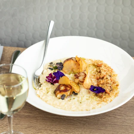 Elegant seafood risotto with scallops and purple flowers, paired with a glass of white wine on a stylish restaurant table.