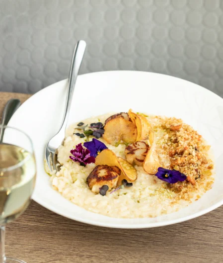 Elegant seafood risotto with scallops and purple flowers, paired with a glass of white wine on a stylish restaurant table.