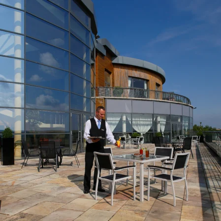 Rooftop terrace with stunning views, modern glass architecture, and a waiter serving drinks at a table.