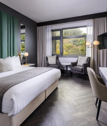 Modern hotel room with king-size bed, stylish decor, large window view, comfy seating, and workspace. Ideal for relaxation.