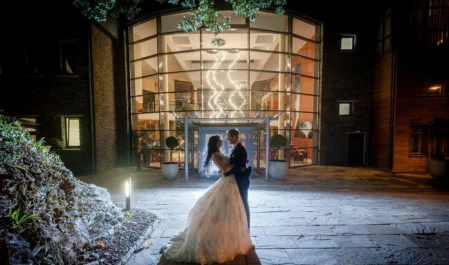 Elegant couple stand illuminated at night in front of luxurious hotel entrance with grand glass façade and modern décor.