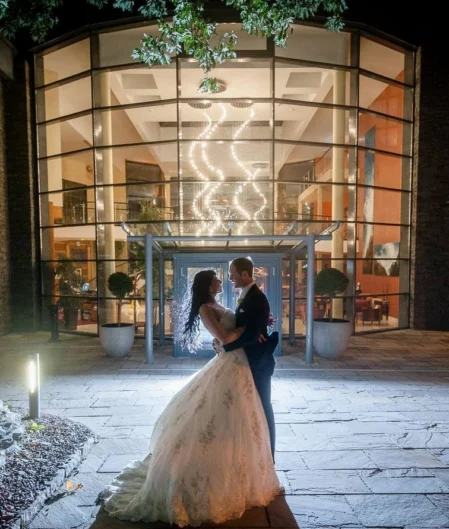 Elegant couple stand illuminated at night in front of luxurious hotel entrance with grand glass façade and modern décor.