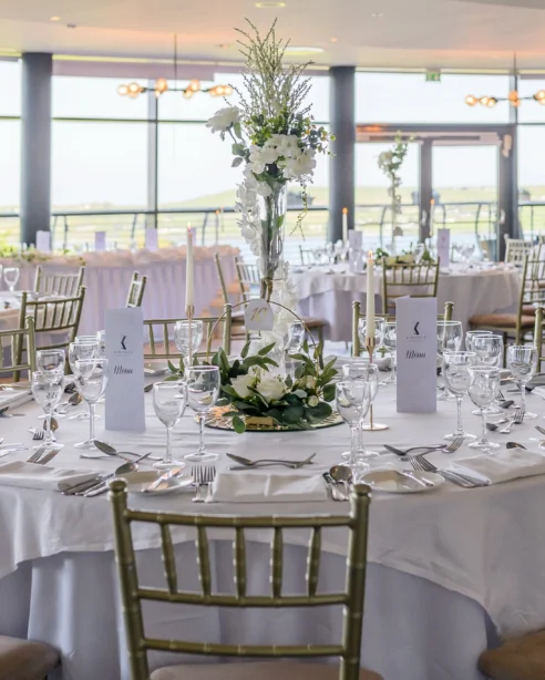 Elegant banquet hall set for a wedding, featuring round tables, white linens, flower centrepieces, and panoramic countryside views.