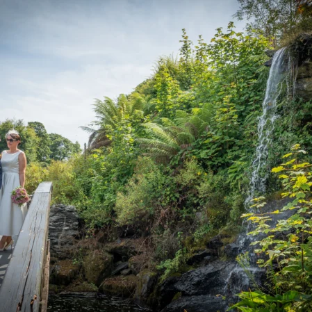 Couple on scenic wooden bridge near lush greenery and waterfall, enjoying serene hotel garden for weddings or relaxation.