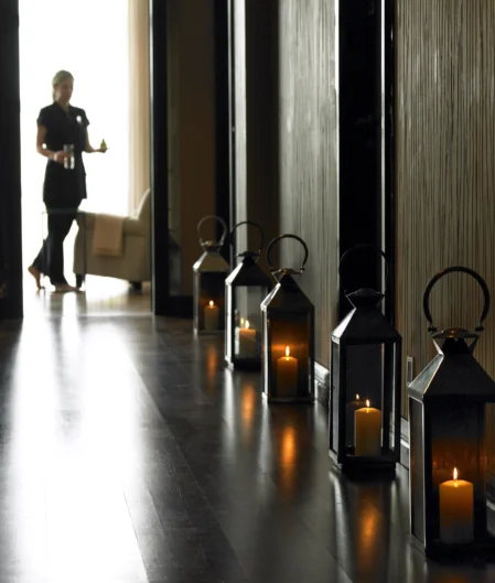 Dimly lit hotel hallway with candle-lit lanterns and a staff member carrying refreshments, leading to a cosy lounge area.