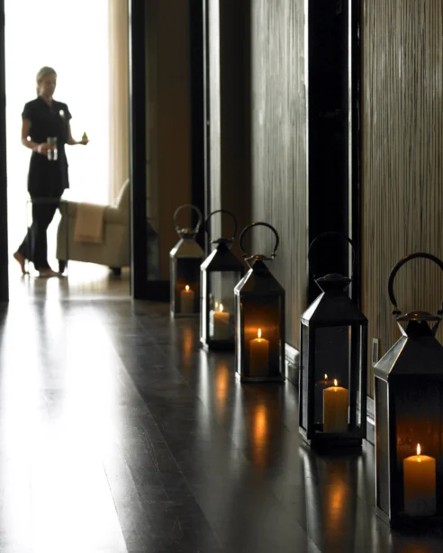 Dimly lit hotel hallway with candle-lit lanterns and a staff member carrying refreshments, leading to a cosy lounge area.